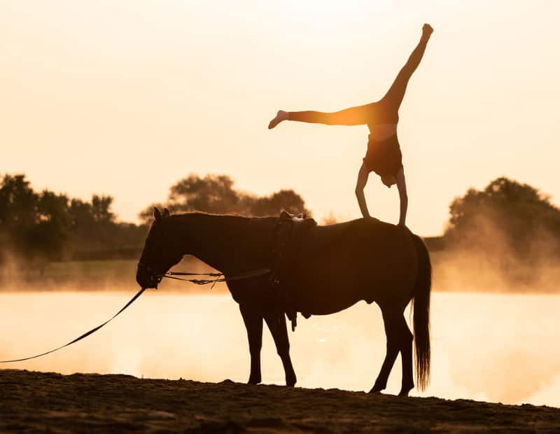 Vaulter doing handstand on horse at sunset beside a river.